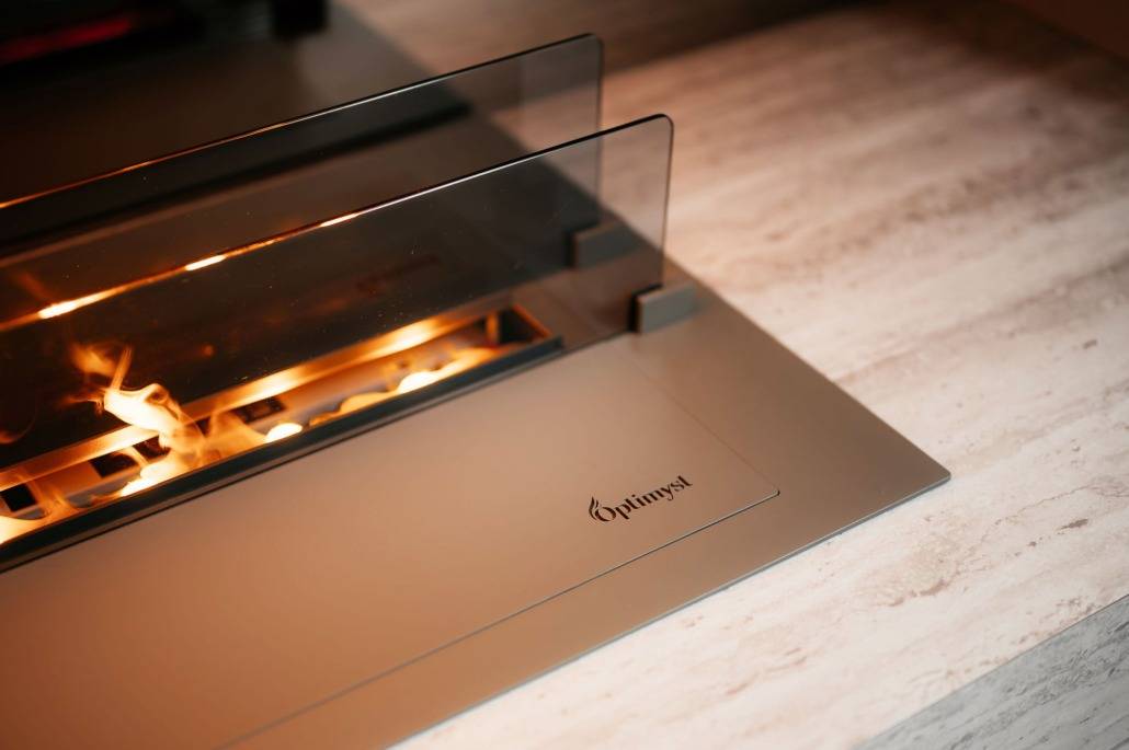 Een moderne tafelhaard met een glazen scherm dat gedeeltelijk een zichtbare vlam omsluit zit op een slank, lichtgekleurd oppervlak - perfect als aanvulling op elke Cinewall opstelling. Het woord Optimyst is afgedrukt op de metalen basis in de buurt van de hoek.