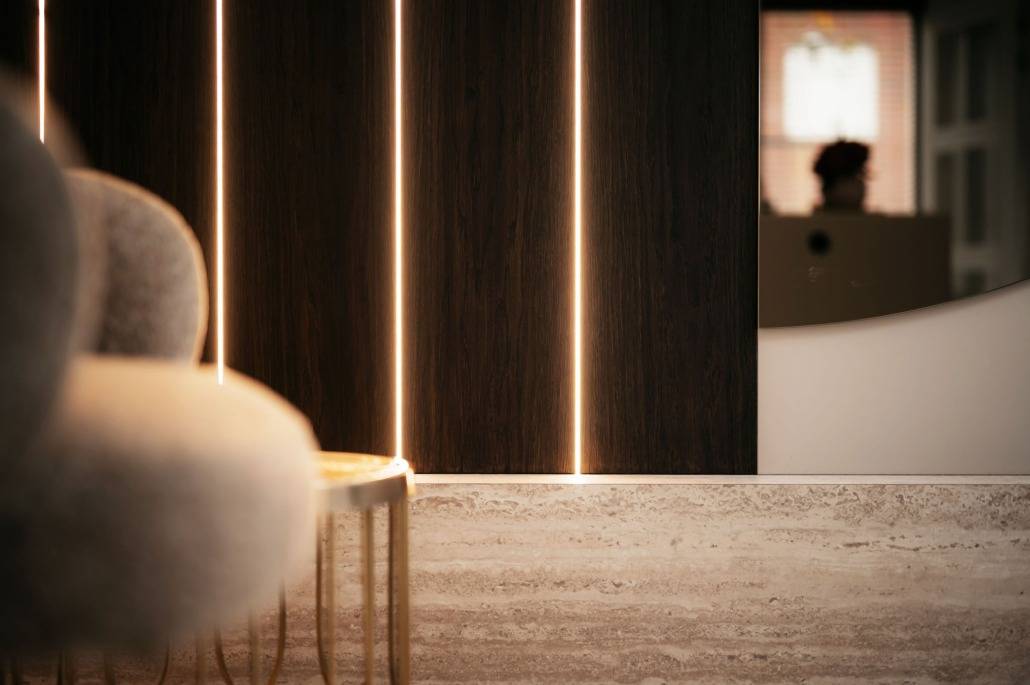 Close-up van een modern interieur met een gebogen grijze stoel, een kleine ronde tafel, een vloer met steentextuur en donkerhouten wandpanelen met verticale LED-lichtstrips en een geïntegreerde cinewall. Een vage reflectie is zichtbaar in een spiegel in de verte.