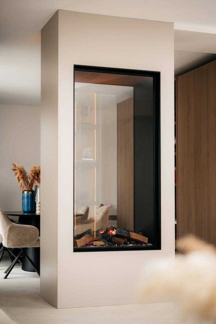 Een moderne, ingebouwde glazen open haard met houtblokken erin zit tussen twee kamers, ontworpen als een cinewall-functie, met neutrale muren en een minimalistische inrichting met gezellige stoelen en een vaas met gedroogd pampagras op de achtergrond.