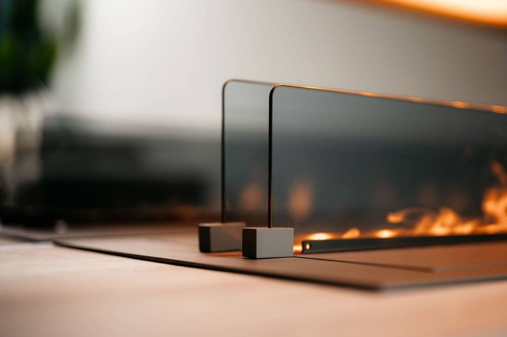 Een close-up van een moderne tafelhaard met glazen panelen en kleine vlammen die binnenin branden, geplaatst op een glad oppervlak in een eigentijdse binnenomgeving naast een strakke cinewall.