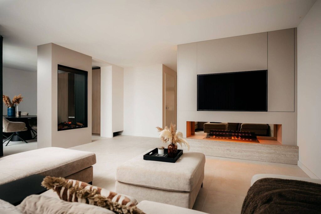Moderne woonkamer met neutrale tinten, met een Cinewall met een tv aan de muur boven een ingebouwde open haard, een grote beige poef, een lichte bank en decoratieve vazen met gedroogde planten. Veel natuurlijk licht vult de minimalistische ruimte.