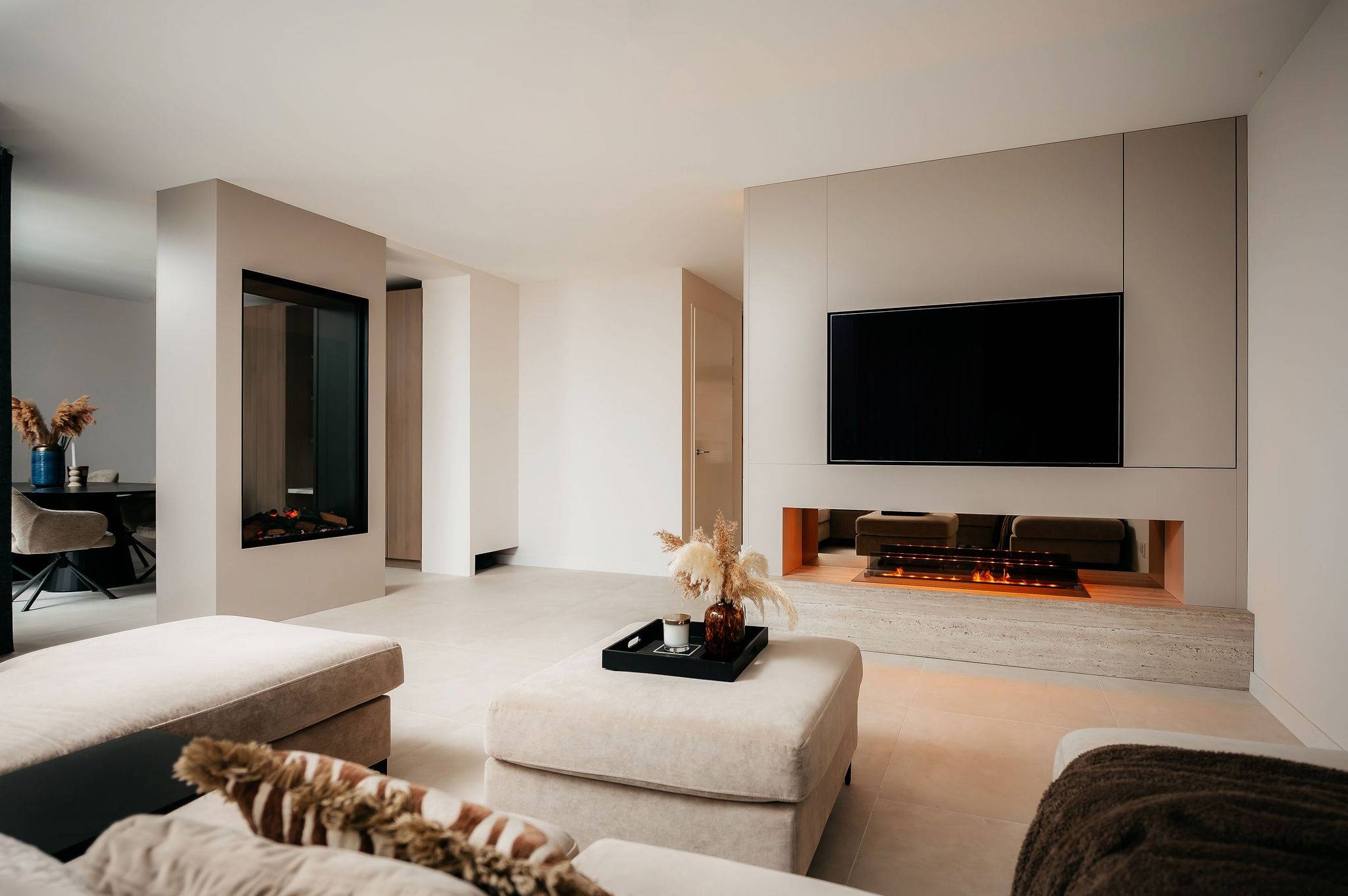 Moderne woonkamer met neutrale tinten, met een Cinewall met een tv aan de muur boven een ingebouwde open haard, een grote beige poef, een lichte bank en decoratieve vazen met gedroogde planten. Veel natuurlijk licht vult de minimalistische ruimte.