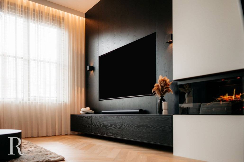 Een moderne woonkamer met een grote flatscreentelevisie op een donkerhouten accentmuur, een lange lage zwarte kast, lichte gordijnen en een open haard met een glazen voorkant. Droog bloemstuk siert de kast.