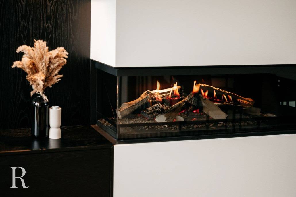 Een moderne, met glas afgesloten open haard met brandende houtblokken, naast een donkere vaas met gedroogd gras en een witte zout- of pepermolen op een strak zwart oppervlak. Het tafereel is minimaal en gezellig.