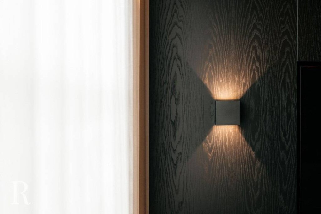 Een moderne wandarmatuur straalt zacht licht uit in een X-patroon op een donkere houten wand, naast een raam bedekt met witte gordijnen.