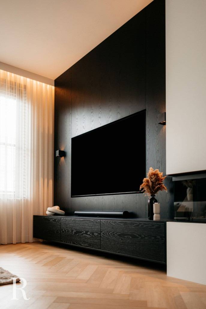 Een moderne woonkamer met een grote flatscreentelevisie aan een donkerhouten accentmuur, een zwarte mediaconsole, een vaas met gedroogde planten, transparante witte gordijnen, een lichthouten visgraatvloer en zacht natuurlijk licht.