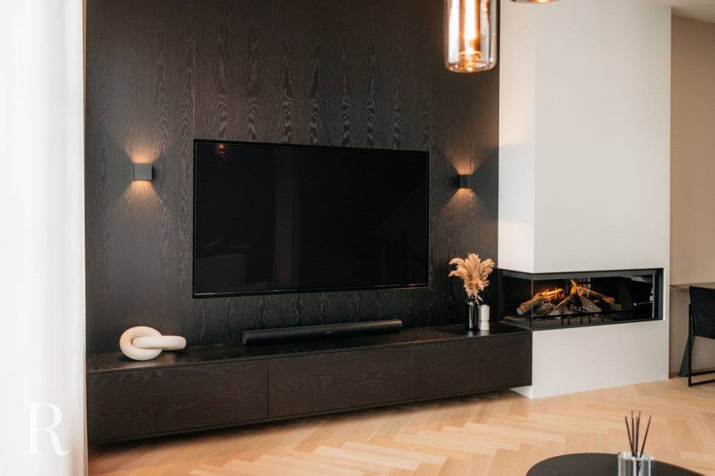 Een moderne woonkamer heeft een grote flatscreentelevisie op een donkerhouten accentmuur, geflankeerd door wandlampen. Hieronder staat een strakke console met decoratieve items en rechts is een ingebouwde open haard zichtbaar.