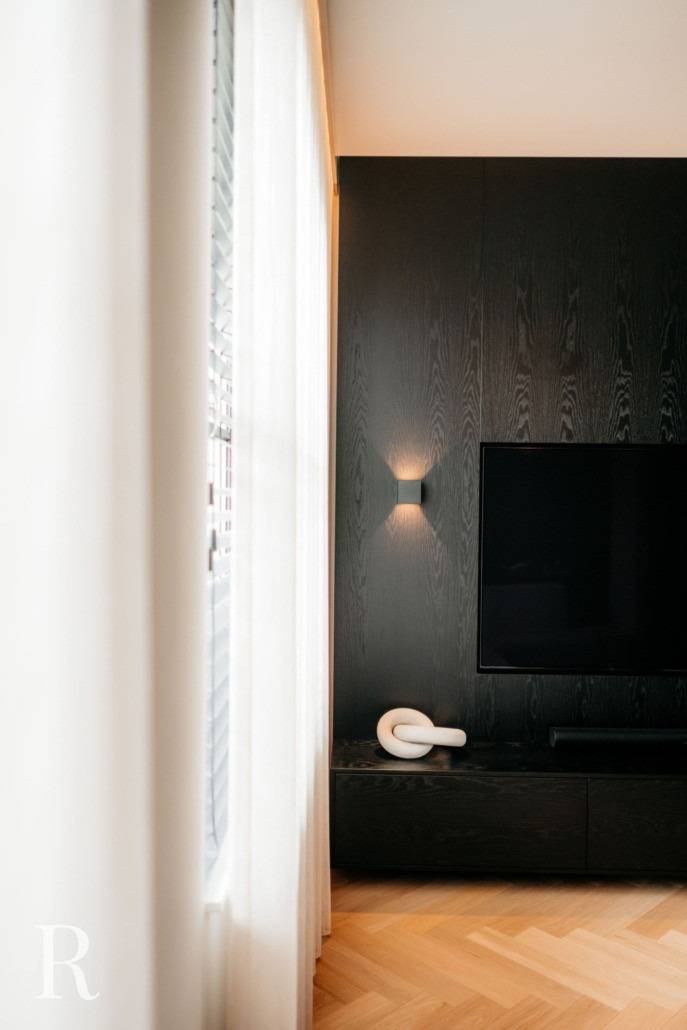 Een moderne woonkamer met een zwarte accentmuur, een gemonteerde flatscreentelevisie, een minimalistisch wit sculpturaal decorstuk op een lage kast en zacht natuurlijk licht dat door transparante gordijnen aan de linkerkant naar binnen valt.