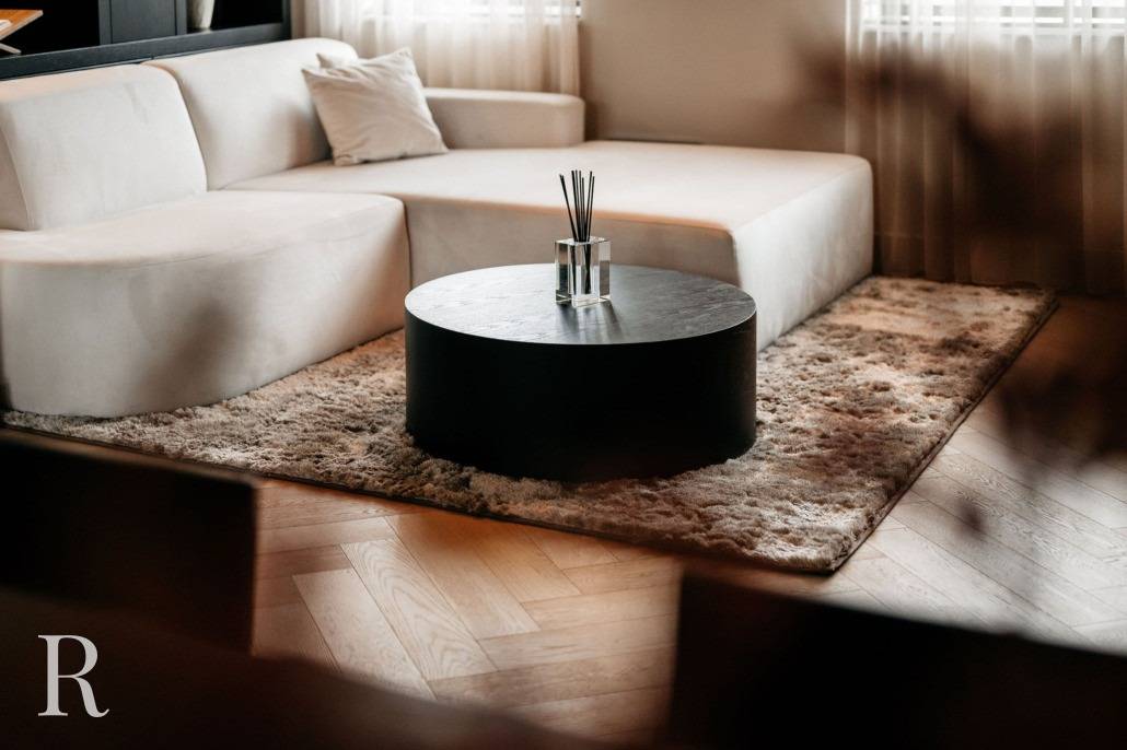 Een moderne woonkamer met een witte sofa, een ronde zwarte salontafel met rieten diffusors, een pluche beige vloerkleed en transparante gordijnen die natuurlijk licht binnenlaten.