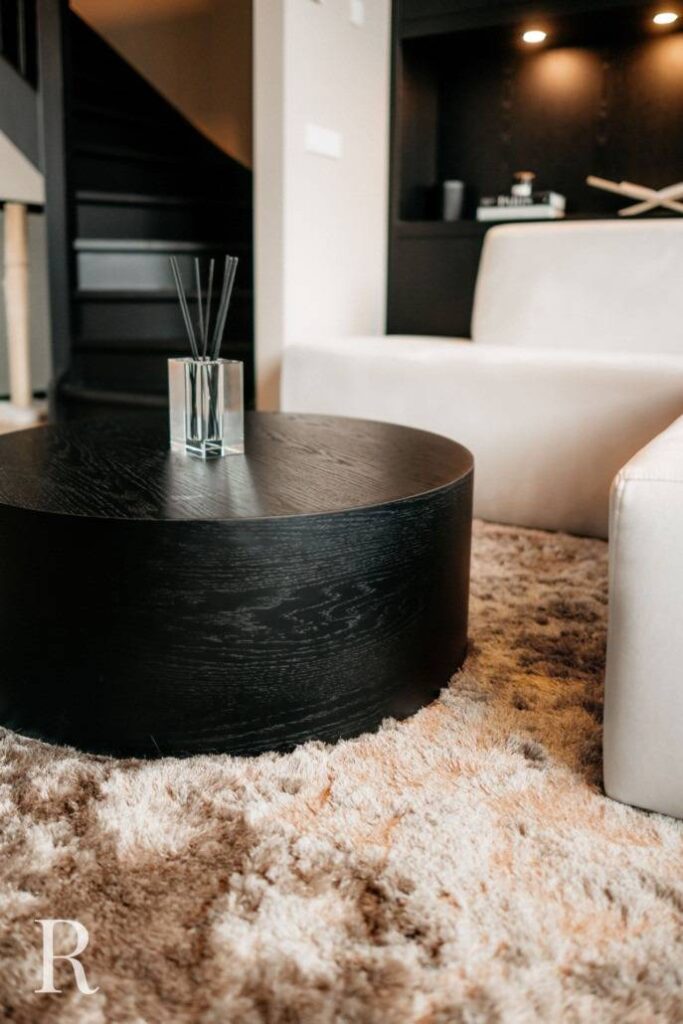 Een moderne woonkamer met een ronde zwarte salontafel op een pluche beige vloerkleed, een glazen rietverdamper op tafel, een witte sofa en een zwarte trap op de achtergrond.