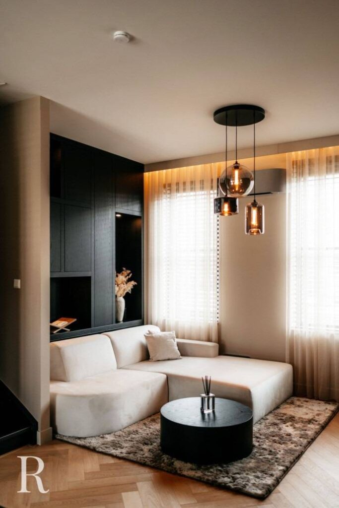 Een moderne woonkamer met een witte sofa, een ronde zwarte salontafel, een vloerkleed met structuur en hanglampen. Vitragegordijnen bedekken de grote ramen en er zijn ingebouwde zwarte planken met decoratieve voorwerpen.