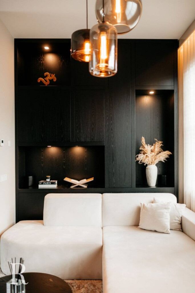 Een moderne woonkamer met een witte sofa, zwarte ingebouwde planken met spots, decoratieve boeken, een vaas met pampagras en hangende amberkleurige glazen hanglampen. Licht stroomt naar binnen vanuit een raam aan de rechterkant.