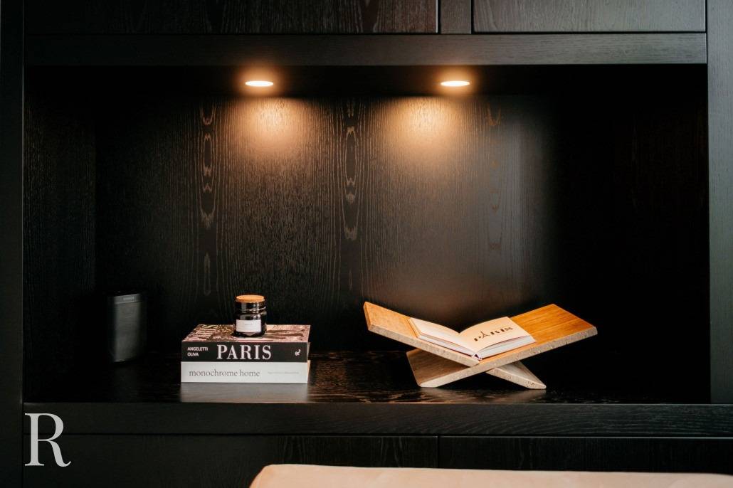 Een zwarte houten plank met twee spots toont een kleine stapel boeken, een kaars en een open boek op een houten standaard. De scène heeft een minimalistische, moderne esthetiek.