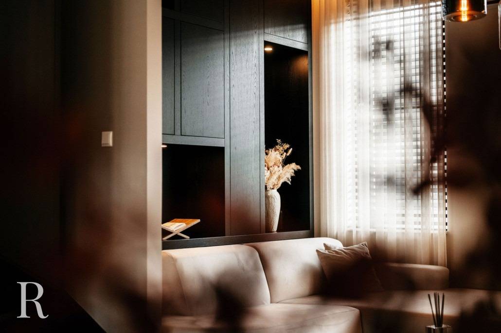 Een moderne woonkamer met een beige bank, een decoratieve vaas met gedroogde bloemen op een donkerhouten plank en zonlicht dat door de transparante gordijnen over een raam valt.