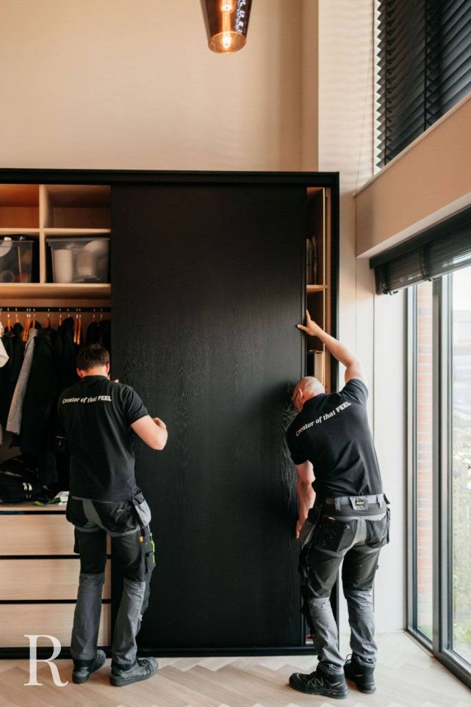 Twee mannen in werkuniformen installeren of verstellen een grote zwarte schuifdeur van een kledingkast in een moderne, goed verlichte kamer met houten vloeren en grote ramen.