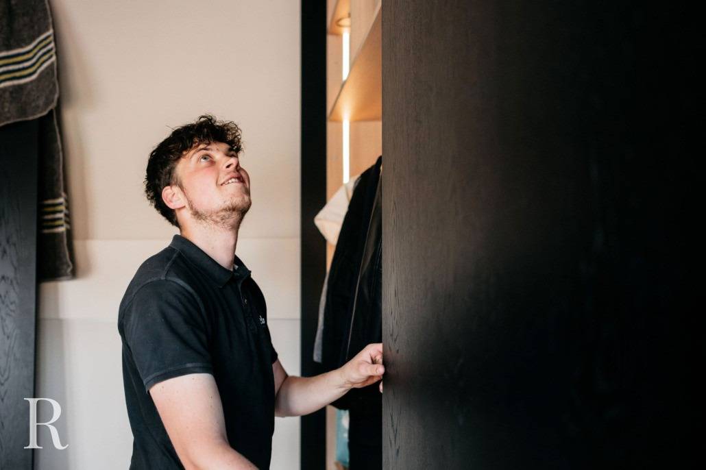 Een man met krullend haar en een korte baard kijkt omhoog terwijl hij bij een open kledingkast staat en naar een plank reikt. Opgevouwen handdoeken zijn zichtbaar aan de linkerkant. De belichting is zacht en natuurlijk.