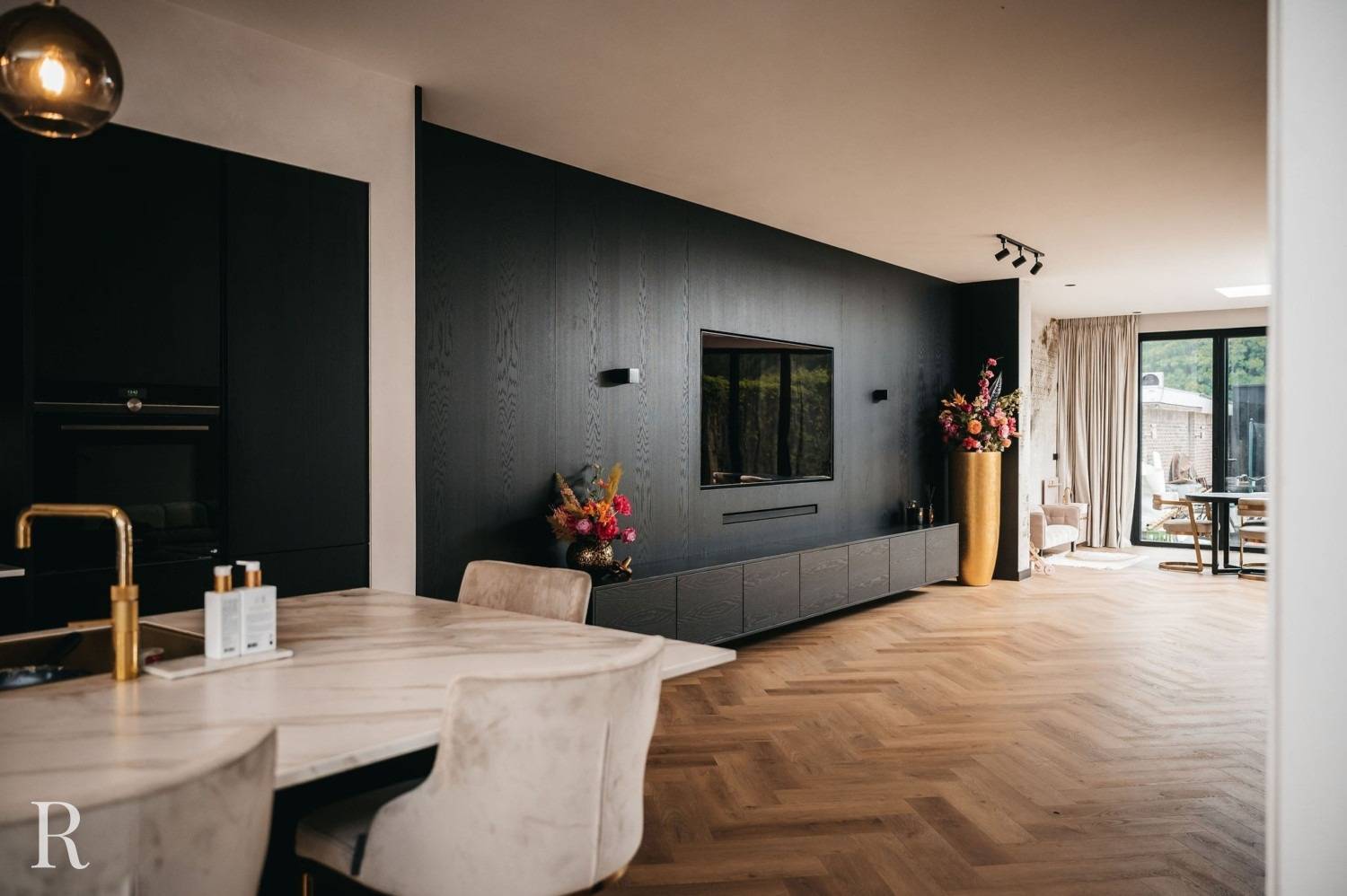 Moderne open woonruimte met visgraat houten vloeren, donkere accentmuur met ingebouwde tv, gouden vaas met kleurrijke bloemen, marmeren eettafel, pluche stoelen en grote ramen die natuurlijk licht binnenlaten.