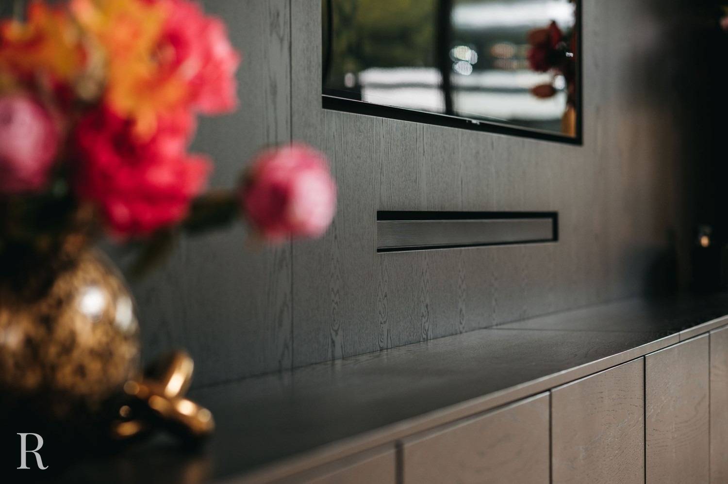 Een close-up van een strak, donker houten kastje met een gemonteerde tv erboven. Een vage vaas met felroze en rode bloemen staat aan de linkerkant van de kast.