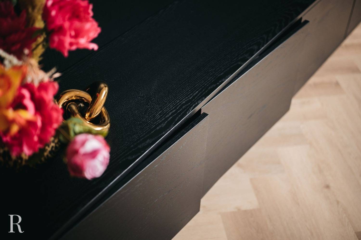 Een close-up van een zwarte houten kast met een modern handvat, naast een vaas met felroze en rode bloemen, op een lichthouten vloer.