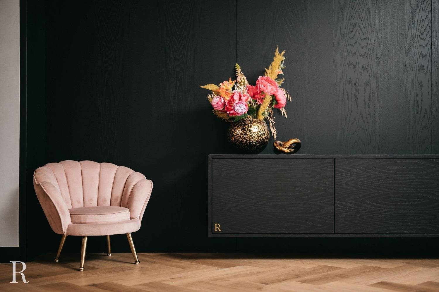 Een roze fluwelen stoel met gouden poten staat naast een zwart wandmeubel met een vaas met felroze bloemen en gedroogd gras; de kamer heeft een lichthouten visgraatvloer en een minimalistisch ontwerp.