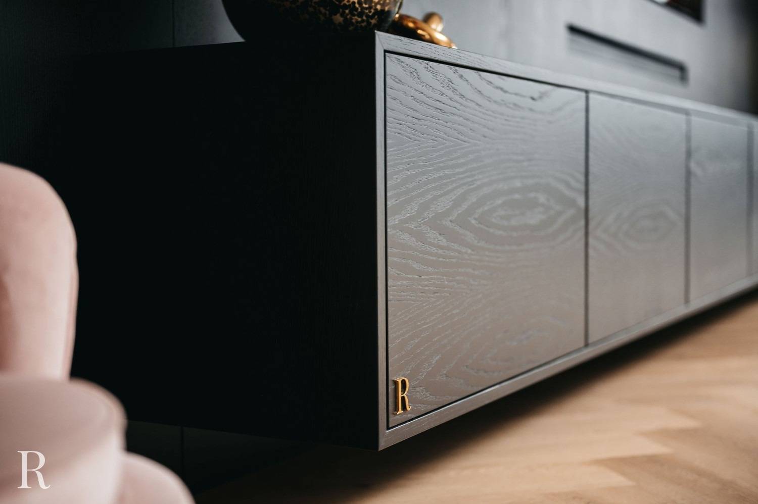 Close-up van een moderne, donkerhouten zwevende kast met een subtiel houtnerfpatroon, gouden accent en een klein R-logo in de hoek, tegen een donkere muur en lichte houten vloer.