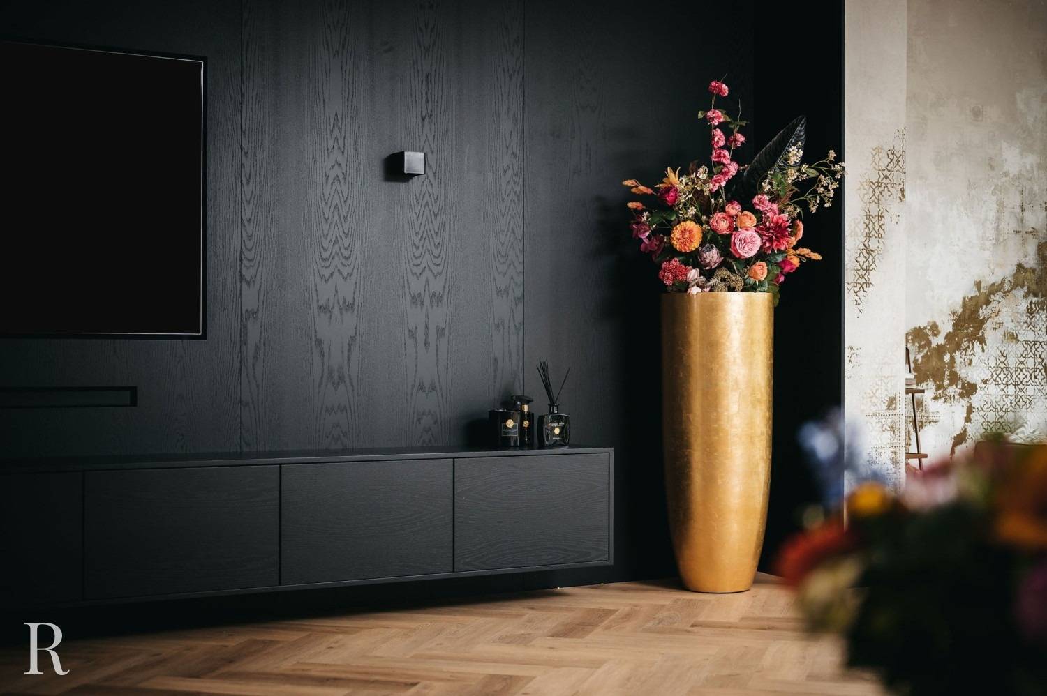 Een moderne woonkamer heeft een donkerhouten mediaconsole en -wand, een gemonteerde tv en een hoge gouden vaas met levendige roze, oranje en rode bloemen op een lichte visgraat houten vloer.