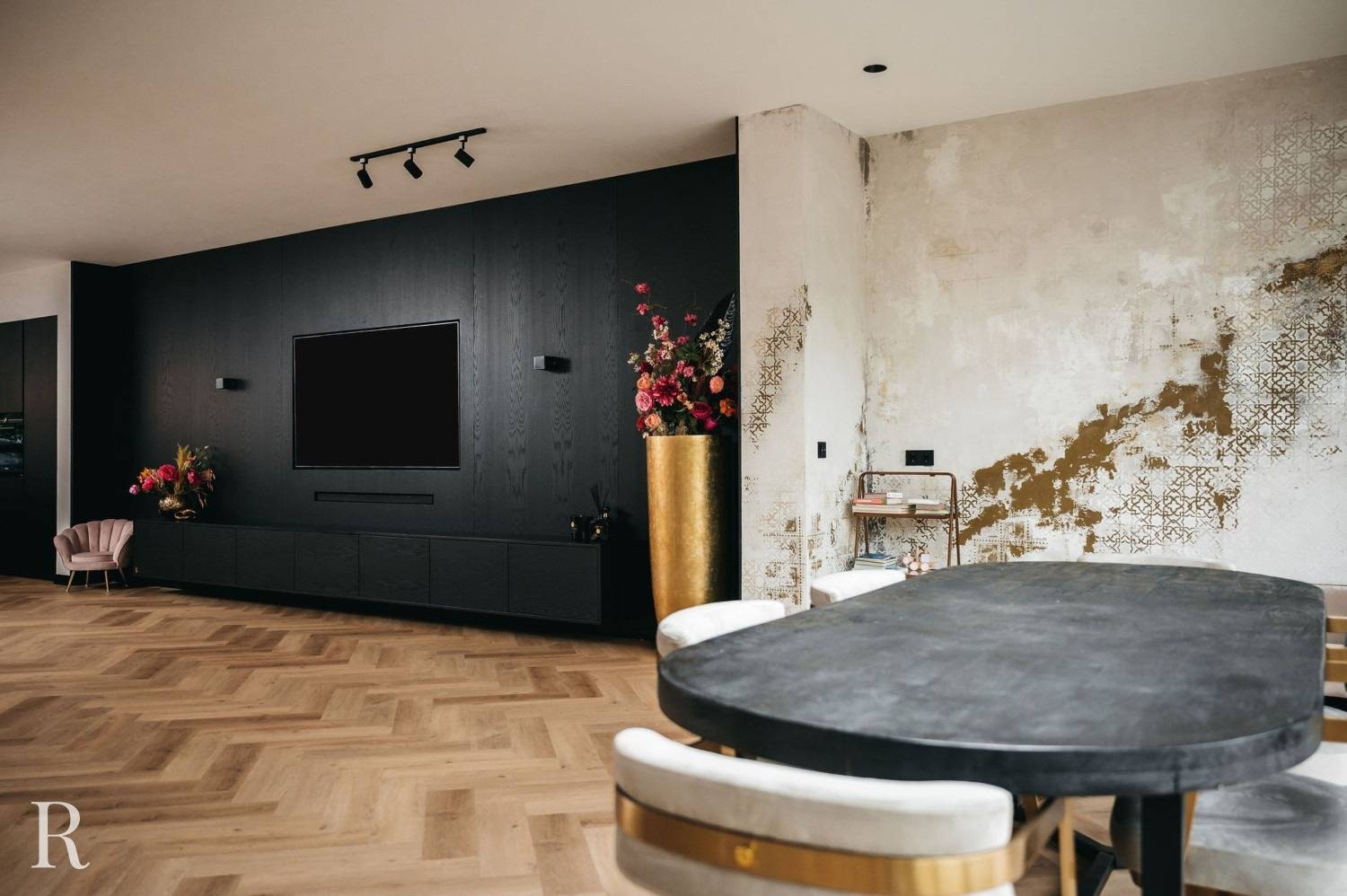 Moderne woon- en eetkamer met visgraat houten vloeren, zwarte accentmuur met tv, gouden vaas met bloemen, witte en gouden accentmuur met structuur en een donkere eettafel met witte en gouden stoelen.