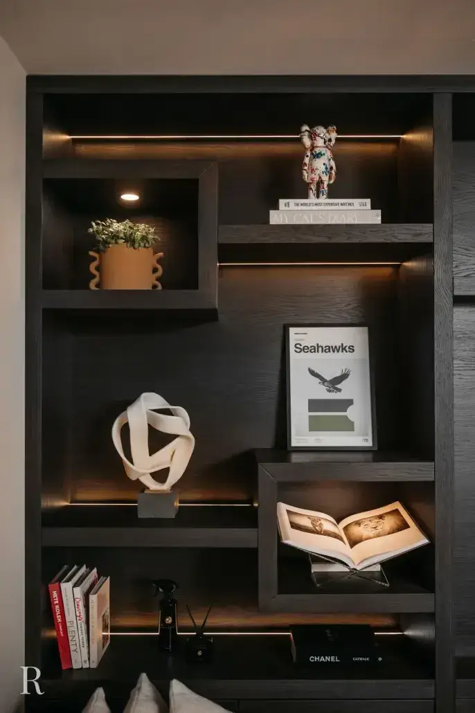 Een moderne zwarte boekenplank met ingebouwde verlichting, met boeken, een potplant, een wit abstract beeldhouwwerk, een Seahawks-print, een Chanel-boek en decoratieve beeldjes op de planken.