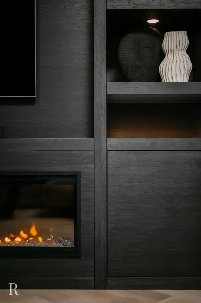 Een modern zwart inbouwmeubel met een glazen schouw aan de linkerkant en twee decoratieve vazen op een plank erboven, de ene zwart met structuur, de andere wit met een golvend ontwerp, zacht verlicht door een kleine spot.