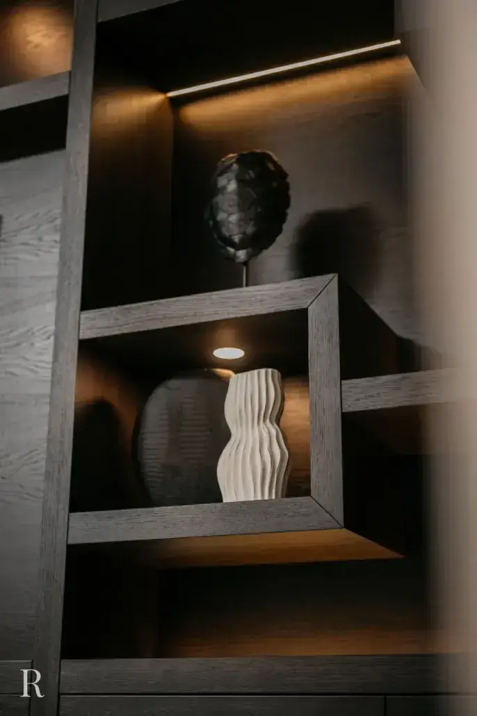 Een moderne houten plank met ingebouwde verlichting toont twee decoratieve objecten: een golvende witte vaas op de voorgrond en een sculptuur met donkere textuur erachter. De verlichting creëert een warme, elegante sfeer.