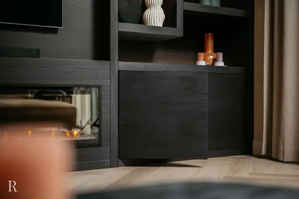 Moderne woonkamer met donkerhouten planken en kasten, decoratieve vazen, kaarsen en een met glas afgesloten open haard. Lichthouten vloeren en beige gordijnen zijn ook zichtbaar.