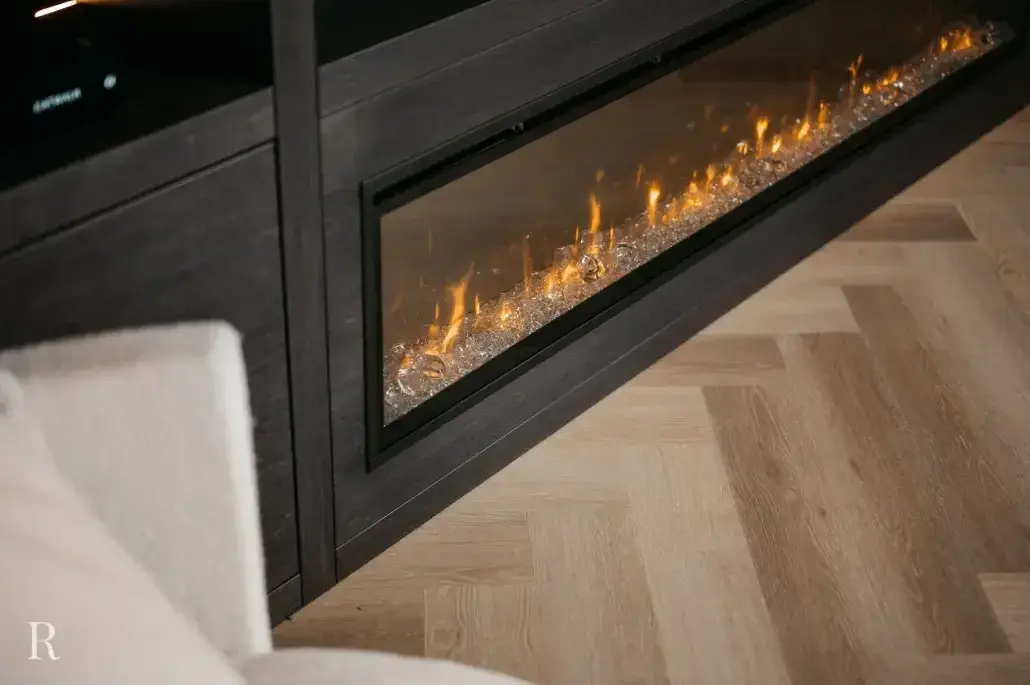 Een moderne lineaire elektrische open haard met gloeiende vlammen en kristallen is ingebouwd in een donkerhouten muur. Op de voorgrond zijn een lichtgekleurde parketvloer en een deel van een lichte stoffen stoel zichtbaar.