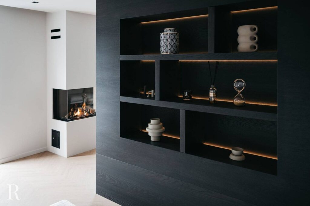 Een moderne leefruimte met een ingebouwde donkerhouten kast met decoratieve items en een minimalistische open haard met glazen voorkant in de hoek. De kamer heeft strakke lijnen en zachte verlichting.
