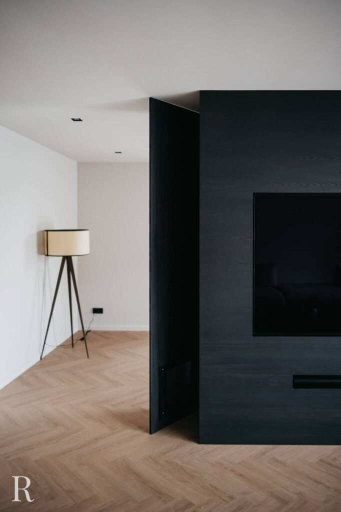 Een minimalistische woonkamer met licht houten visgraatvloer, een modern zwart wandmeubel met een tv en een driepoot vloerlamp met een rechthoekige kap in de hoek.