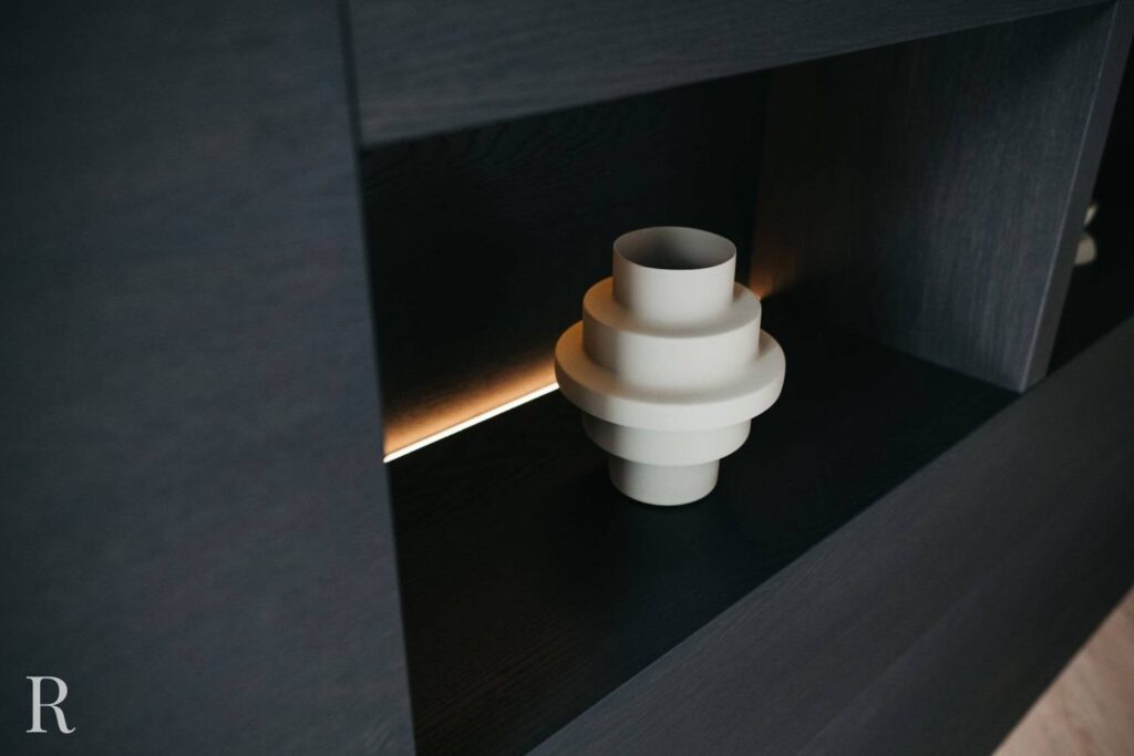 Een moderne, geometrische witte vaas staat op een donkere houten plank, met warm tegenlicht dat het gelaagde ontwerp benadrukt. In de hoek van de afbeelding is een vaag R-watermerk te zien.