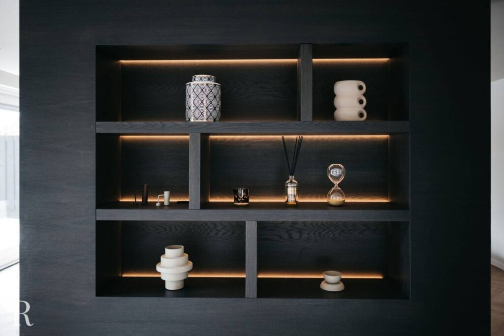Een donkere houten stelling met ingebouwde verlichting toont decoratieve voorwerpen, waaronder vazen, een zandloper, een pot met patroon, diffusors en moderne keramische beelden.
