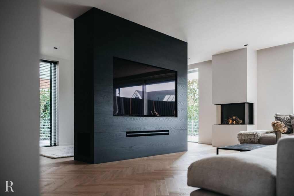 Moderne woonkamer met een grote zwarte entertainmentmuur met een gemonteerde tv, een open haard in de hoek, neutrale meubels, een visgraatvloer en grote ramen die natuurlijk licht binnenlaten.