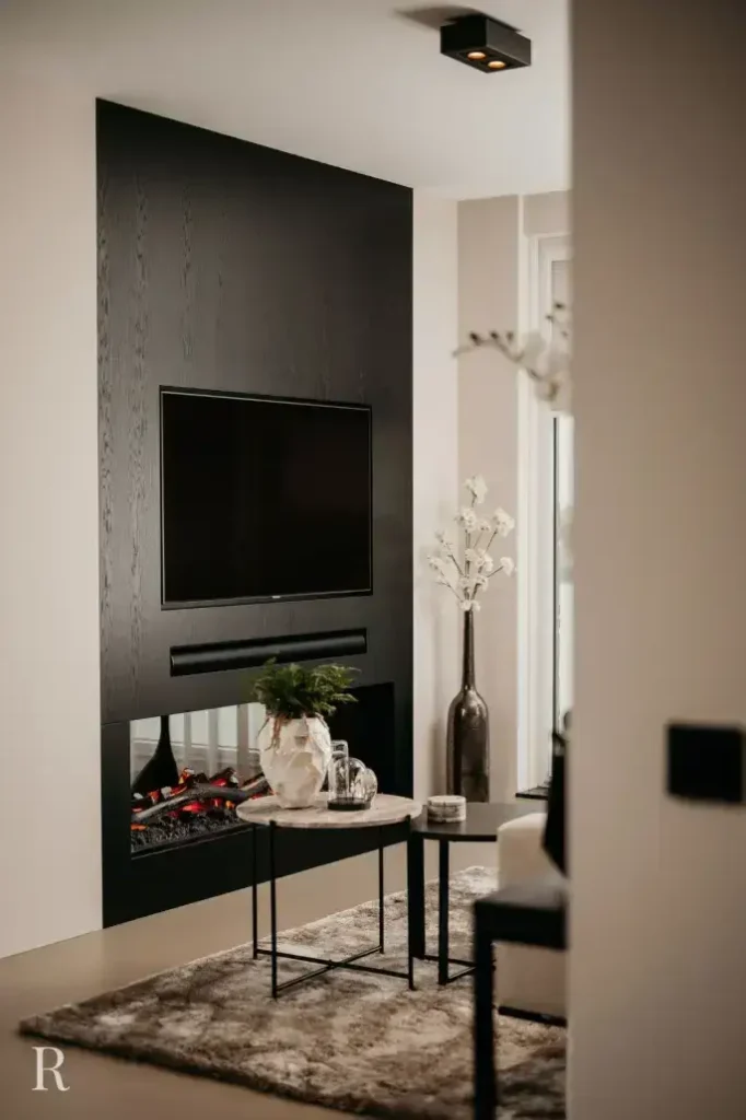 Een moderne woonkamer met een strakke zwarte muur met een gemonteerde tv en ingebouwde elektrische open haard, een marmeren salontafel met planten en decor, en een zacht grijs vloerkleed op de vloer.