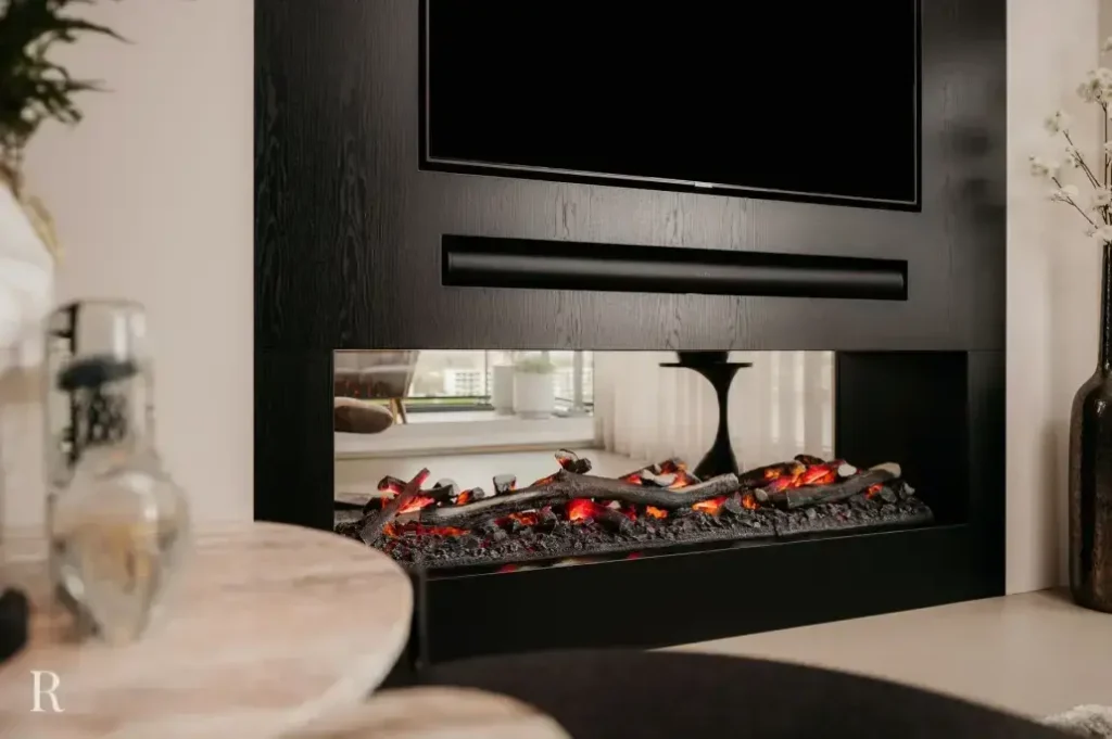 Een moderne elektrische open haard met gloeiende houtblokken is ingebouwd in een strak zwart wandmeubel onder een gemonteerde tv. De kamer heeft een neutrale inrichting, een ronde tafel en decoratieve vazen.