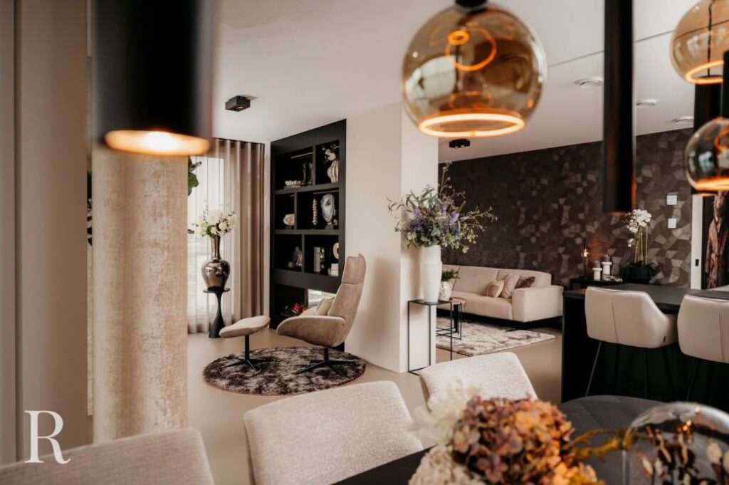 Een moderne, elegante woon- en eetkamer met neutrale tinten, stijlvolle verlichting, een gezellige fauteuil, decoratieve vazen, bloemstukken en een accentmuur met patroon op de achtergrond.