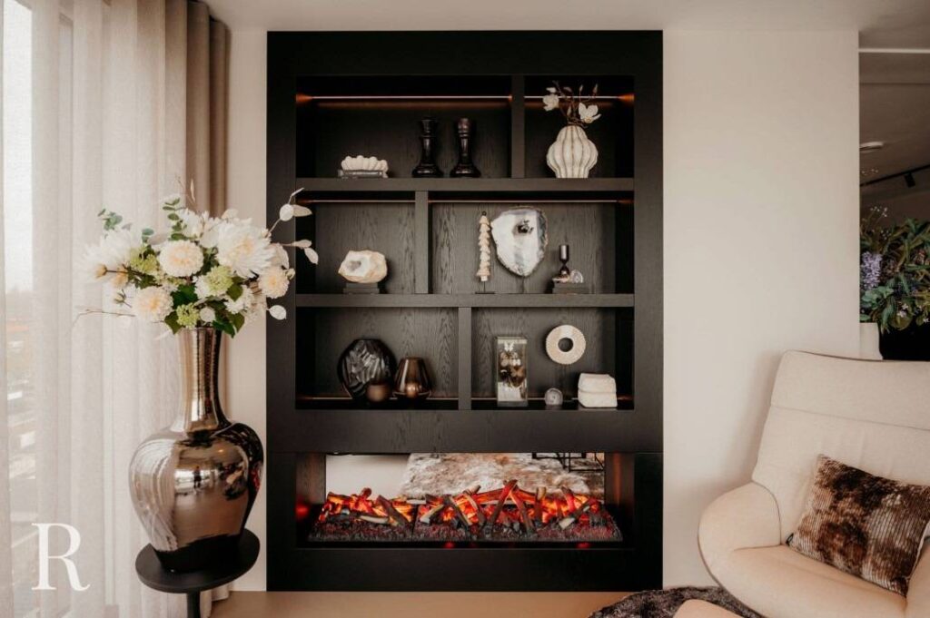 Een modern zwart rek met decoratieve objecten en kunstwerken staat boven een elektrische open haard. Een grote vaas met witte bloemen staat links naast een gezellige beige fauteuil met kussen in een stijlvolle woonkamer.
