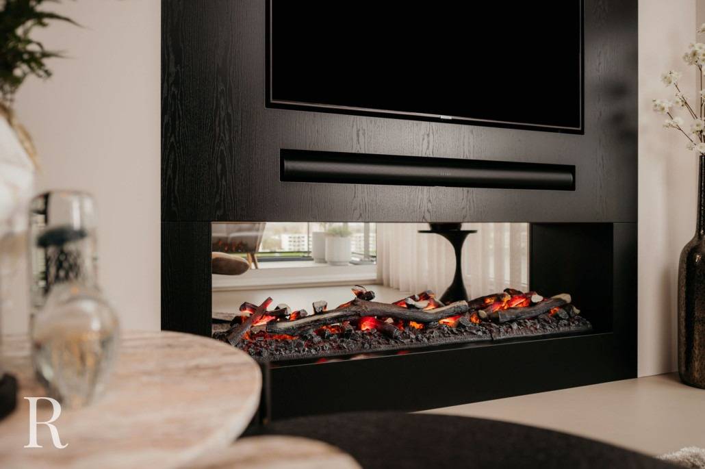 Moderne woonkamer met een strakke zwarte elektrische open haard onder een tv aan de muur, decoratieve houtblokken en gloeiende sintels, een ronde marmeren tafel op de voorgrond en stijlvolle inrichting op de achtergrond.