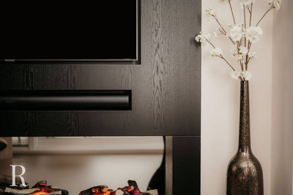 Een moderne woonkamer met een zwarte houten open haard, een tv erboven, een soundbar onder de tv, een decoratieve vaas met witte bloemen rechts en imitatie haardhout met gloeiende sintels onderaan.