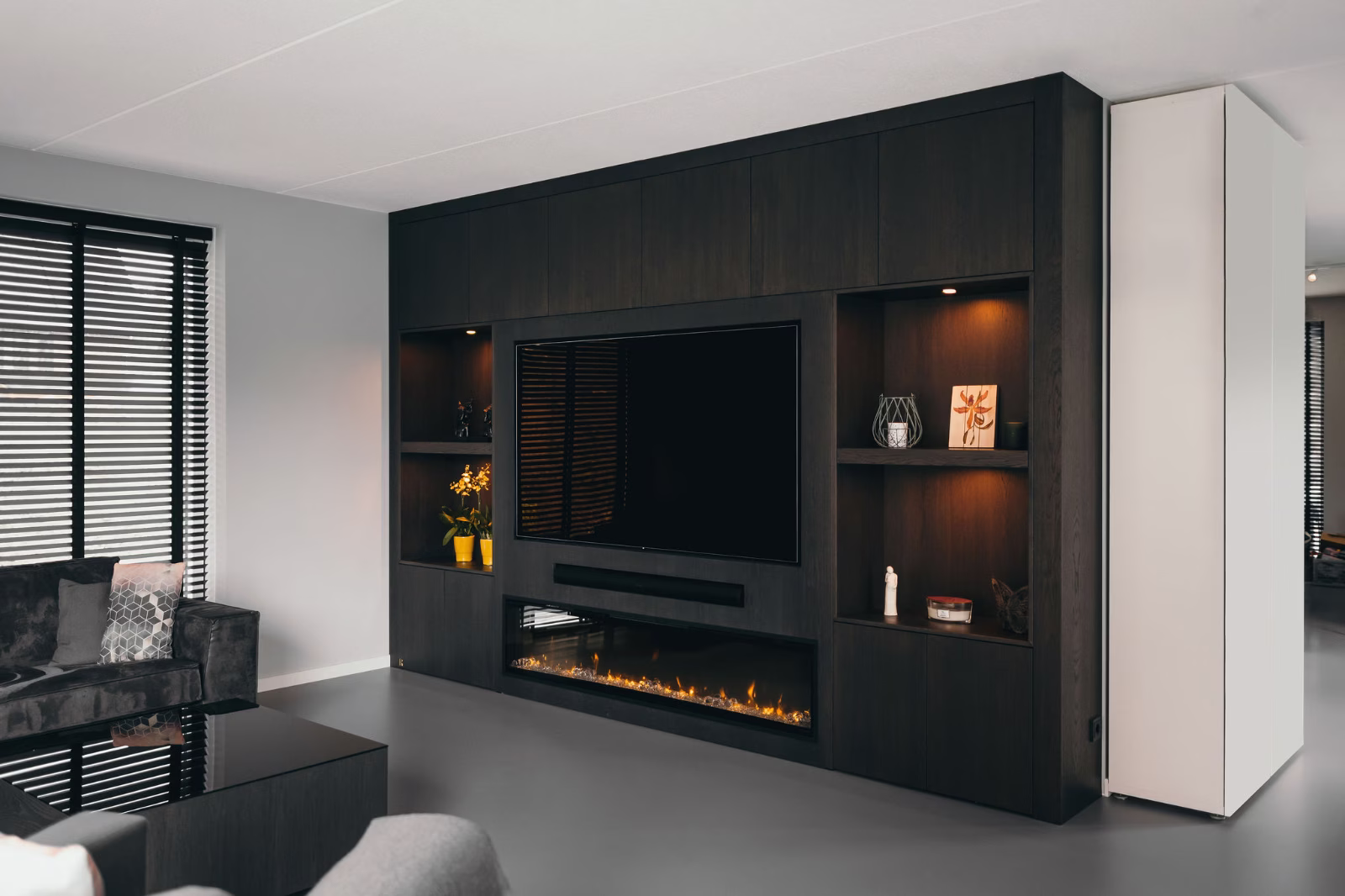 Een moderne woonkamer heeft een donker ingebouwd entertainmentcentrum met een grote tv, planken met decoraties en een elektrische open haard eronder. Een glazen salontafel en een grijze bank zijn ook zichtbaar.