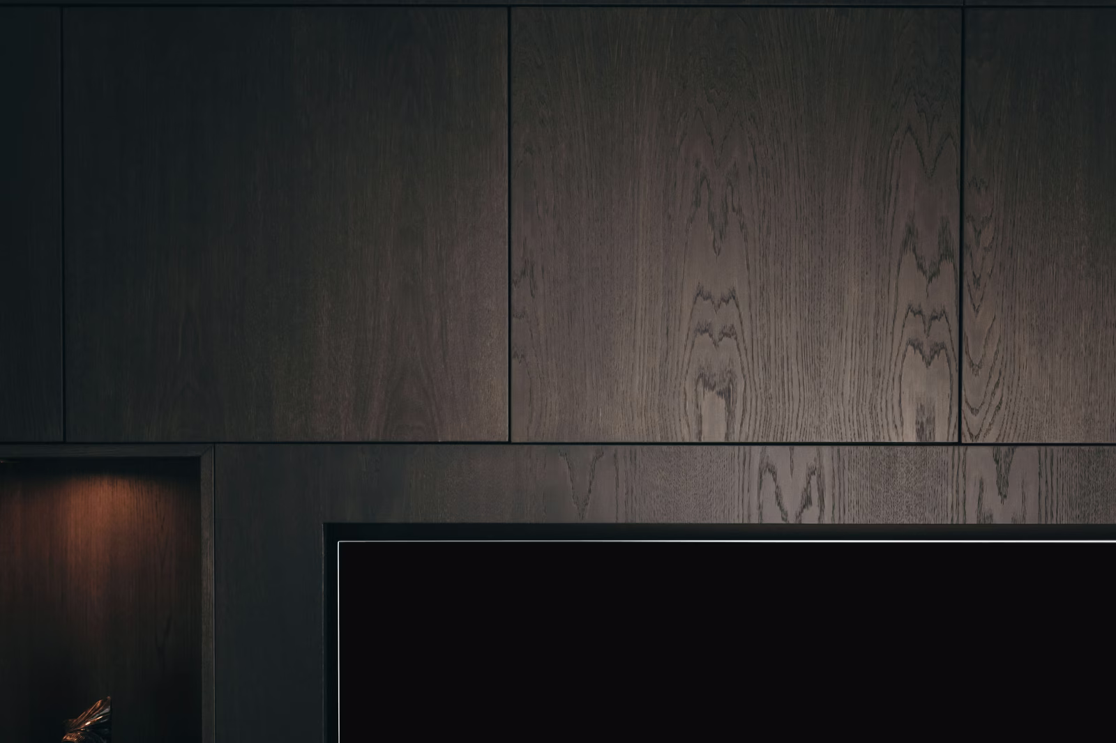 Donkerhouten kasten met een gladde, matte afwerking worden getoond boven de bovenrand van een televisiescherm; een kleine ingebouwde plank met een zwak licht is gedeeltelijk zichtbaar aan de linkerkant.