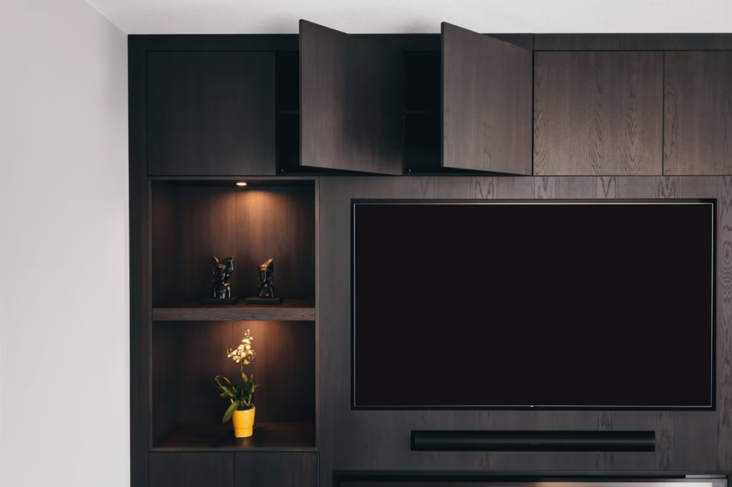 Een modern donkerhouten entertainmentmeubel met een grote tv, twee open bovenkasten, een plank met twee zwarte kattenbeeldjes en een onderste plank met een potplant in een gele pot, allemaal verlicht door ingebouwde verlichting.