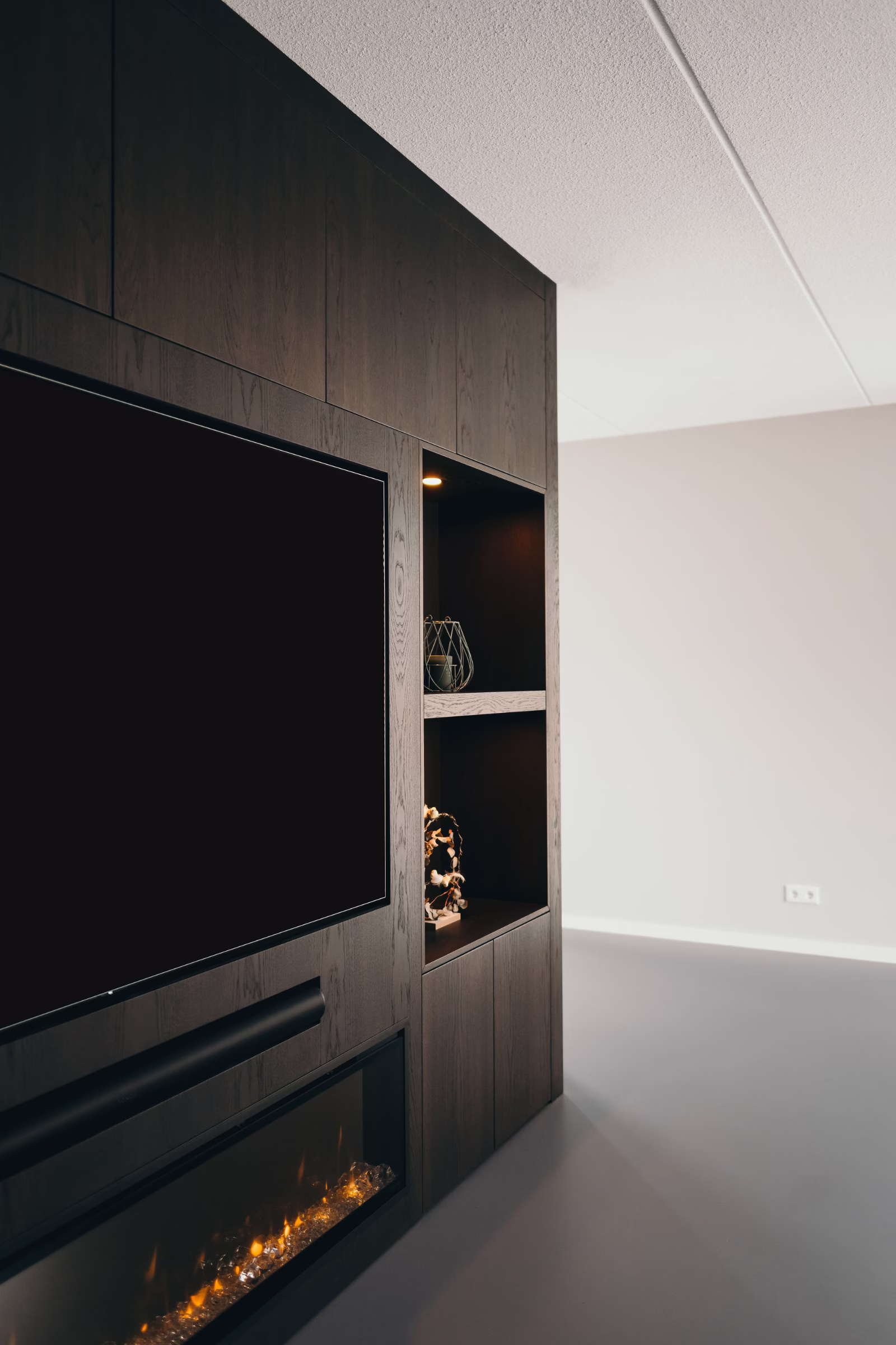 Een moderne woonkamer met donkerhouten ingebouwde planken, een flatscreentelevisie, een elektrische open haard en decoratieve voorwerpen tegen een lichtgekleurde muur en grijze vloer.