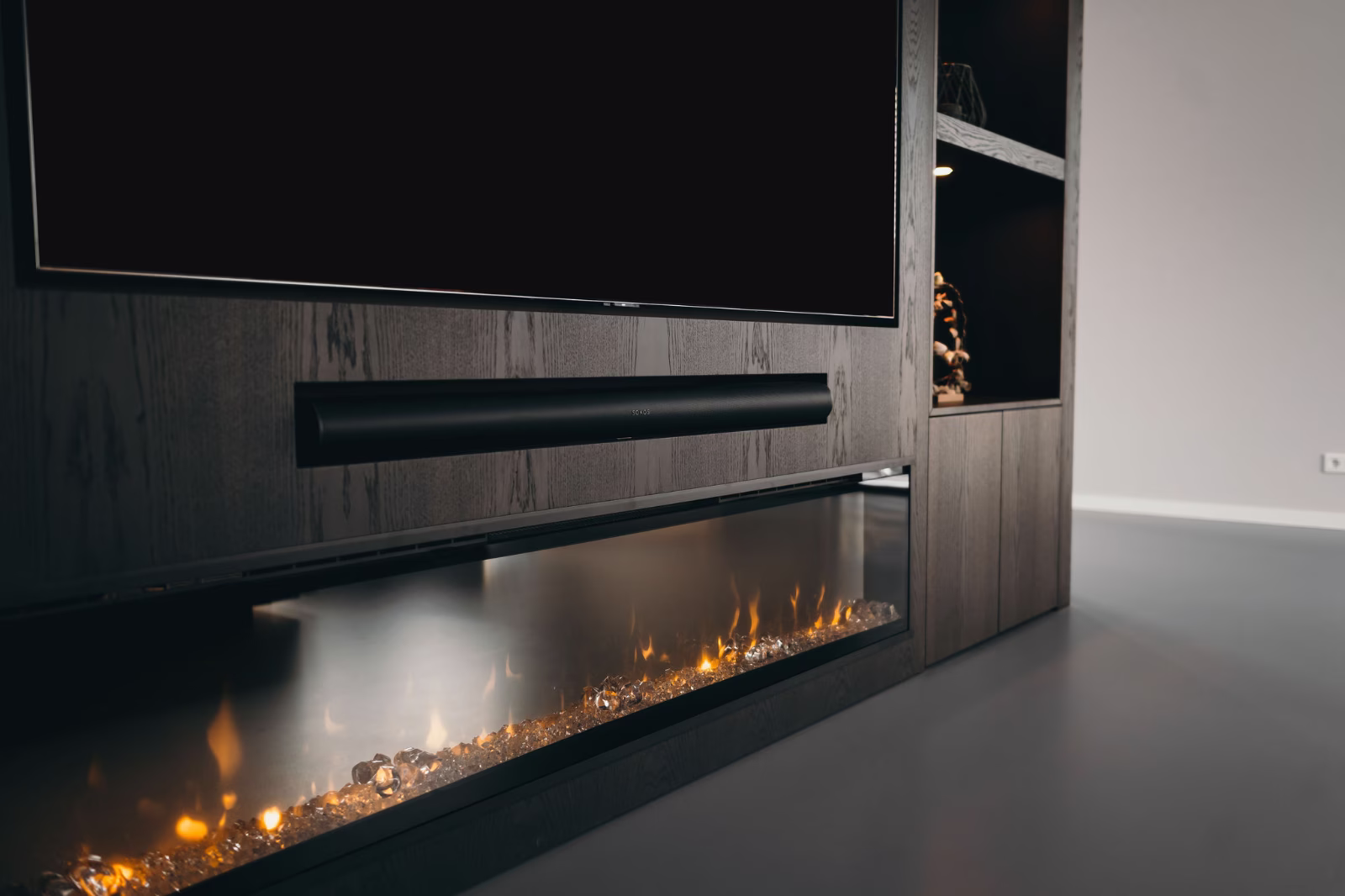 Een moderne woonkamer met een flatscreen-tv aan de muur, een slanke zwarte soundbar eronder en een elektrische open haard met gloeiende vlammen en kristallen decoraties eronder, allemaal in een slanke houten entertainmentkast.