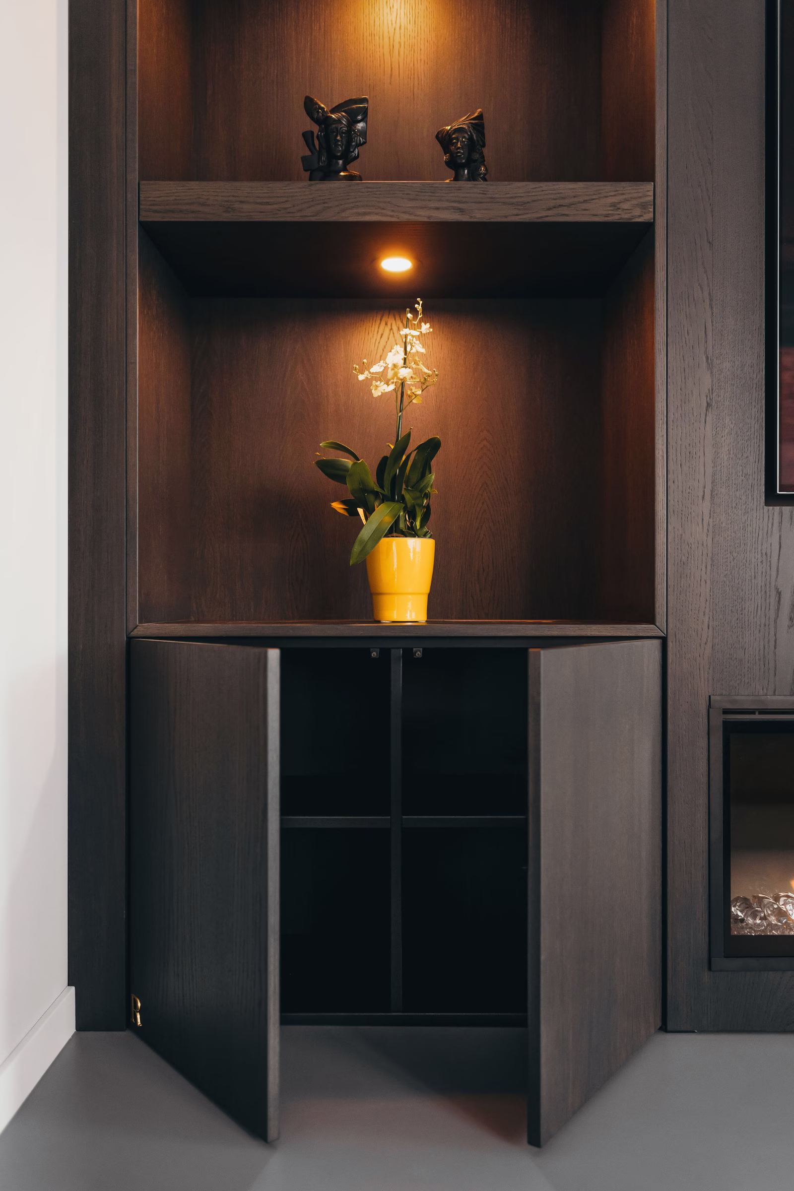 Een houten kast met open onderste deuren onthult lege planken. Op de middelste plank staat een gele pot met een bloeiende plant onder een spot, en op de bovenste plank staan twee decoratieve voorwerpen.