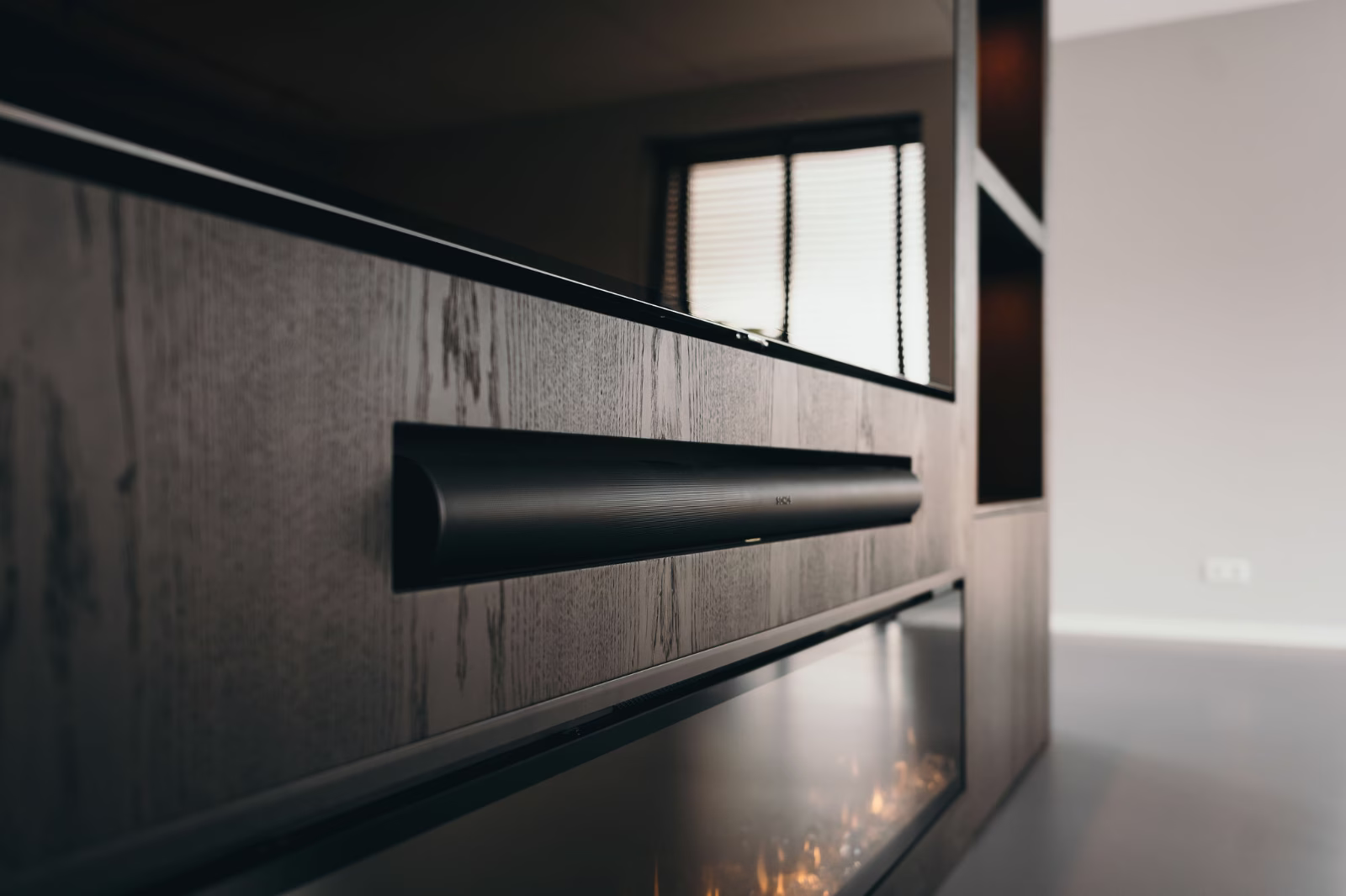 Een close-up van een moderne zwarte soundbar gemonteerd op een houten entertainmentmeubel, met een flatscreen-tv erboven en een raam met jaloezieën op de onscherpe achtergrond.